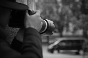 Osoba w kapeluszu trzymająca aparat fotograficzny z teleobiektywem, obserwująca z ukrycia, w tle samochód. Detektyw Warszawa.