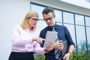 Dwoje pracowników biurowych w okularach stojących na zewnątrz budynku – kobieta w różowej koszuli wskazuje palcem na tablet trzymany przez mężczyznę w granatowej koszuli z krawatem